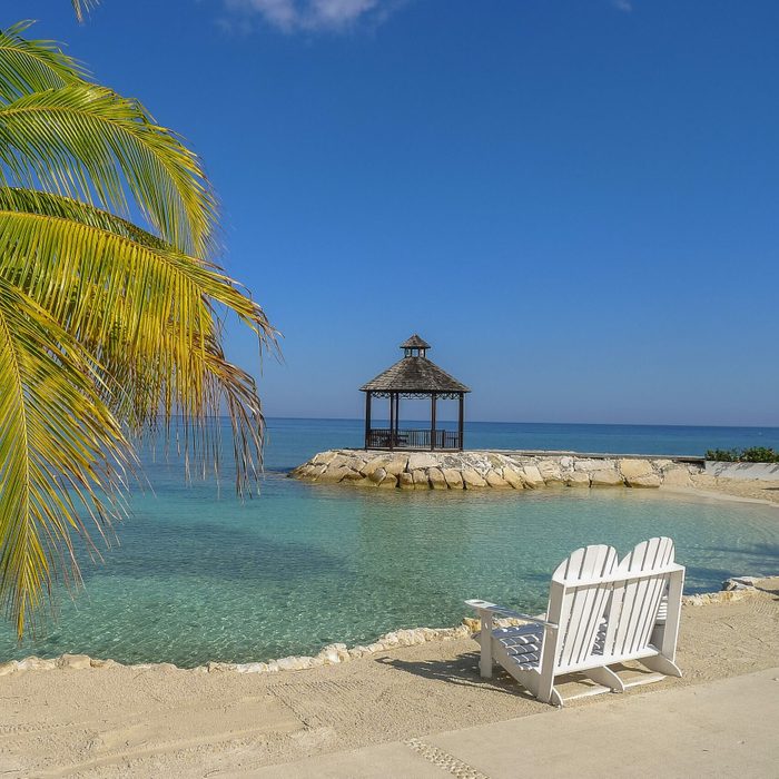 Palm tree shades beach chairs facing a gazebo over clear blue water; everything bathed in bright sunlight under a clear sky.