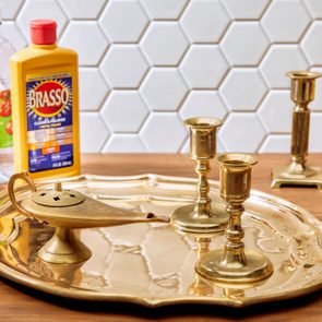 Brass objects rest on a polished tray, surrounded by cleaning items, in front of a white hexagonal-tile wall.