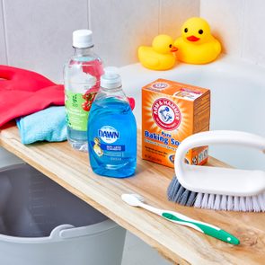 Cleaning supplies rest on a wooden board over a bathtub, including gloves, vinegar, soap, baking soda, scrub brush, and a toothbrush. Duck toys float nearby.