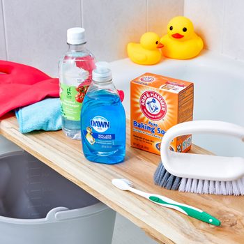 Cleaning supplies rest on a wooden board over a bathtub, including gloves, vinegar, soap, baking soda, scrub brush, and a toothbrush. Duck toys float nearby.