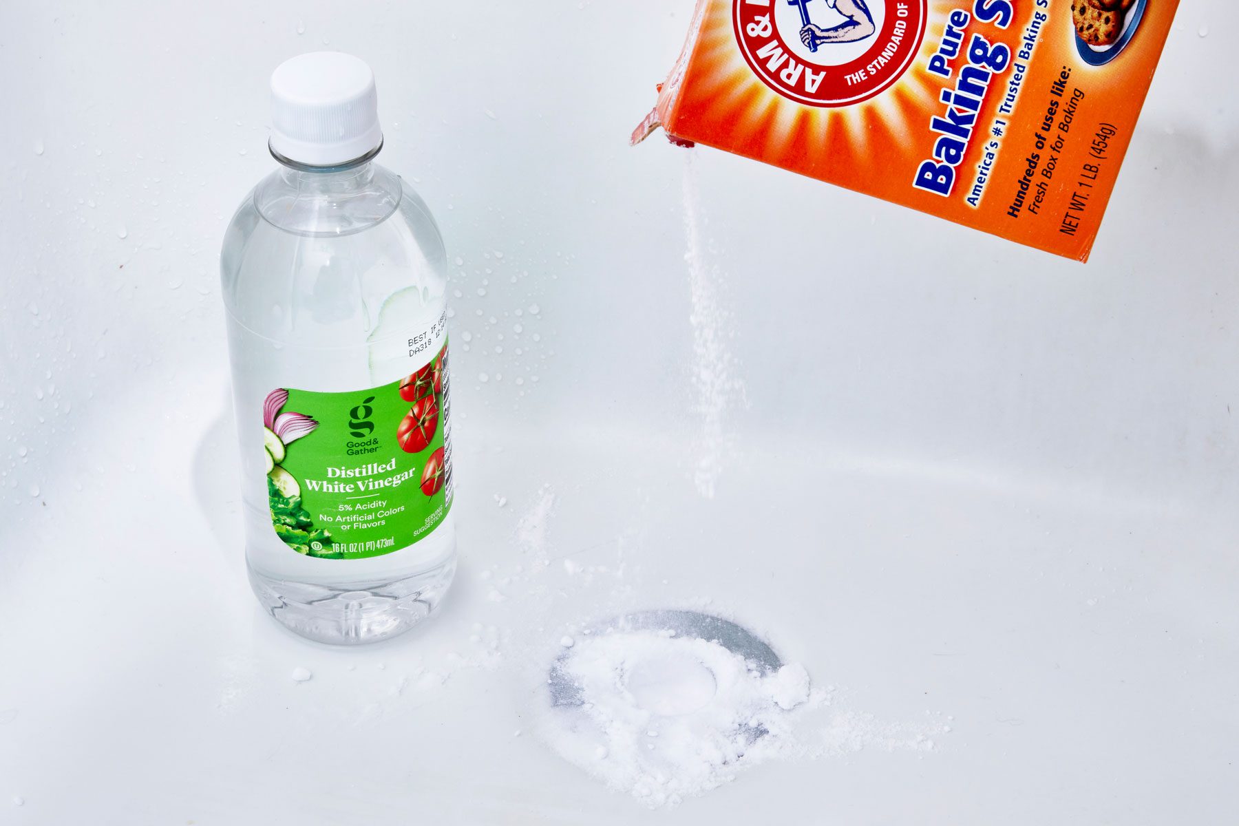 pouring baking soda into a bathtub drain, bottle of white vinegar nearby