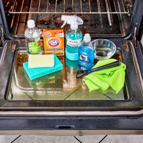 Cleaning supplies, including sponges and sprays, displayed on an open oven door in a kitchen setting. Items: vinegar, baking soda, dish soap, rag, scraper.