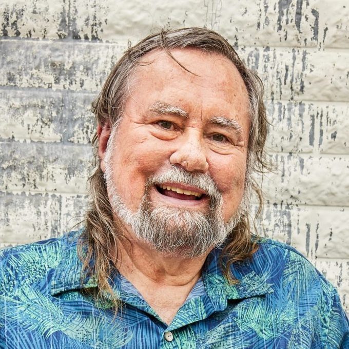 A smiling man with long hair and a beard is wearing a blue patterned shirt, standing against a textured, lightly painted brick wall.