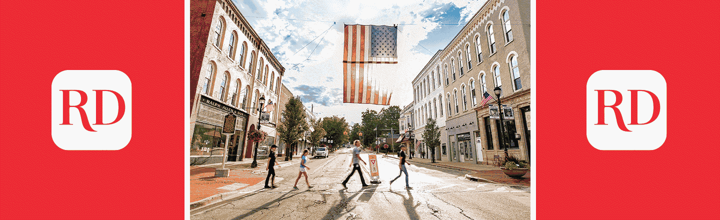 People crossing a street under a hanging American flag in a small town, surrounded by historic brick buildings and trees, with a blue sky above.