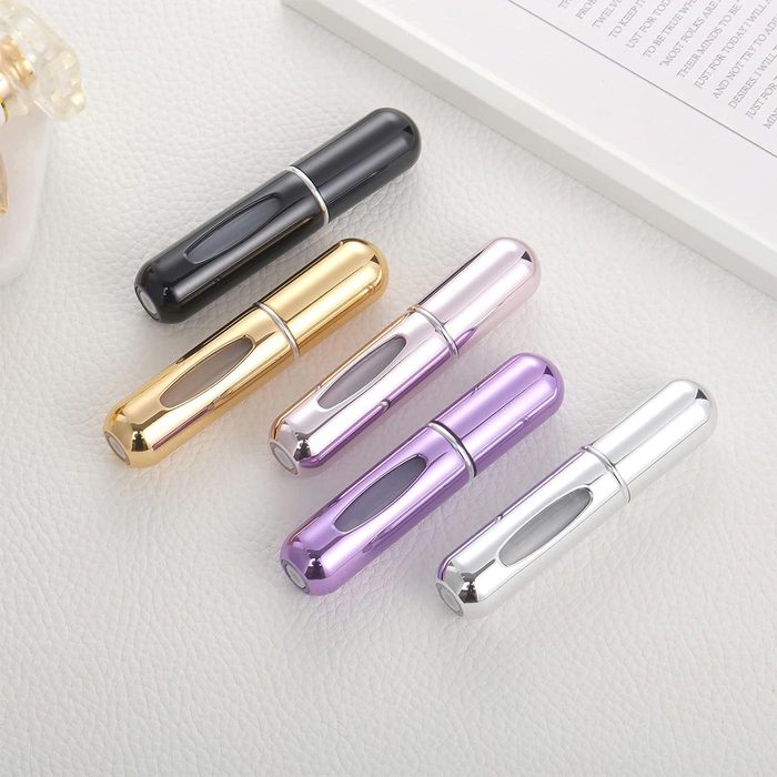 Five metallic perfume atomizers rest diagonally on textured white leather. They are colored black, gold, pink, purple, and silver.