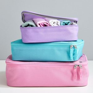Stacked packing cubes, zipped, containing rolled clothes, are set against a plain gray wall on a white surface.
