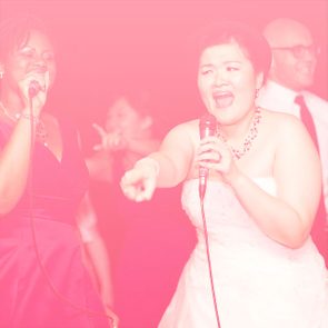 People sing energetically into microphones, surrounded by others in a celebratory, pink-tinted atmosphere.