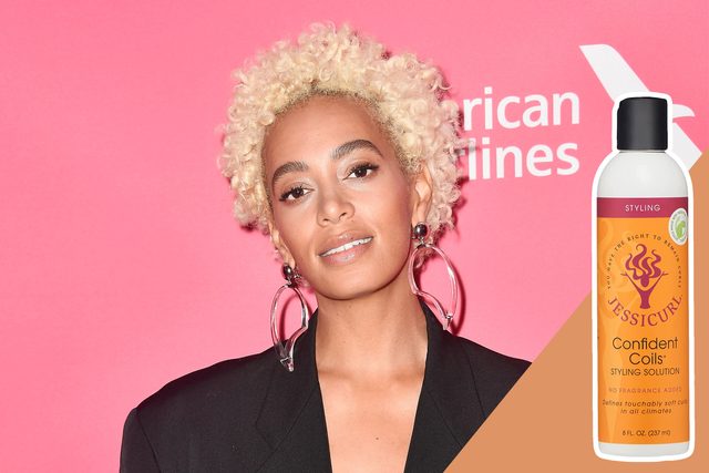 A person poses against a pink backdrop. They wear large hoop earrings. A bottle of 