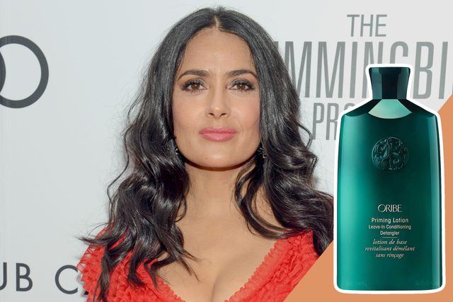 A woman stands confidently, wearing a red dress, in front of a wall with text, next to an Oribe Priming Lotion bottle.