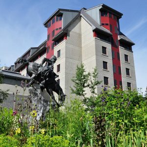 Statue of a rider on horseback leaping, surrounded by colorful flowers and greenery, with a tall building in the background under a blue sky.