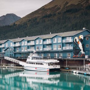 Boat docked beside blue building with puffin mural; forested mountains in the background.