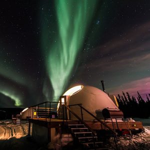 Domed igloo-like structures sit illuminated under vivid green Northern Lights in a snowy, starry landscape with silhouetted trees.
