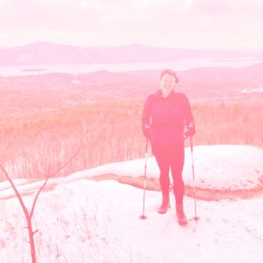 A person stands smiling, holding trekking poles on a snowy hilltop, with a vast forest and mountains in the background.
