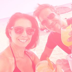 Two people wearing sunglasses smile while holding drinks, sitting on a sandy beach under sunny skies.