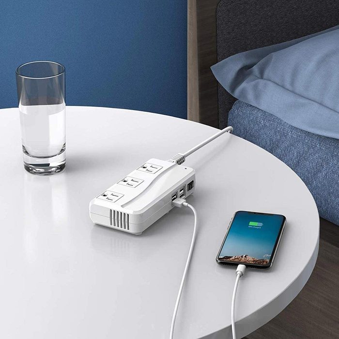 Power strip charges a smartphone on a white table beside a glass of water, near a bed with blue bedding.