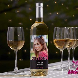 Wine bottle displays a woman's photo; text reads, â€œ20 + ONE, Drink up B*TCHES! Love, Lilly.â€ Surrounded by four filled glasses outdoors.