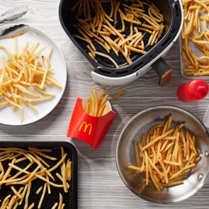 Fries are being served on a table with an air fryer, baking sheet, pan, plates, and McDonald's packaging around.