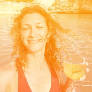 Person holds a wine glass, smiling, standing near a body of water with sunlit cliffs in the background.