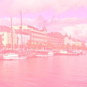 Boats dock calmly beside a waterfront lined with historic buildings. Sky is partly cloudy, casting a pinkish filter over the scenic harbor view.