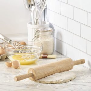 Rolling pin rests on dough, surrounded by a whisk in a bowl, egg shells, and flour jar on a tiled kitchen counter.