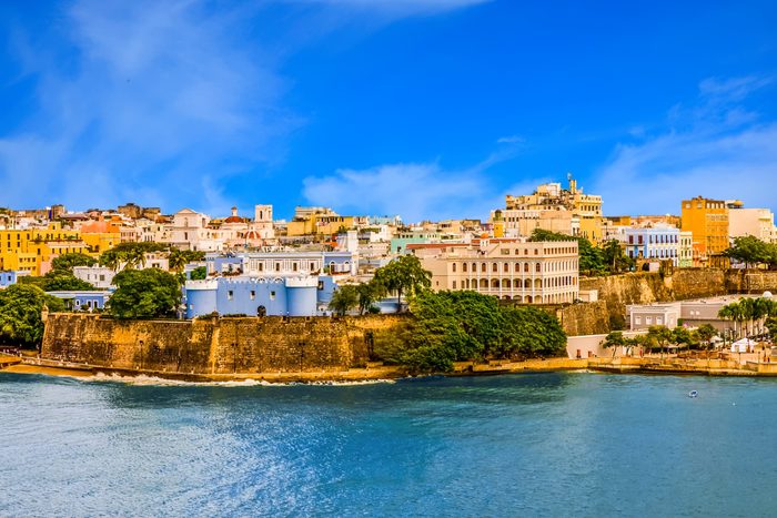 Colorful buildings sit atop ancient fort walls, overlooking the blue coastline under a clear sky.