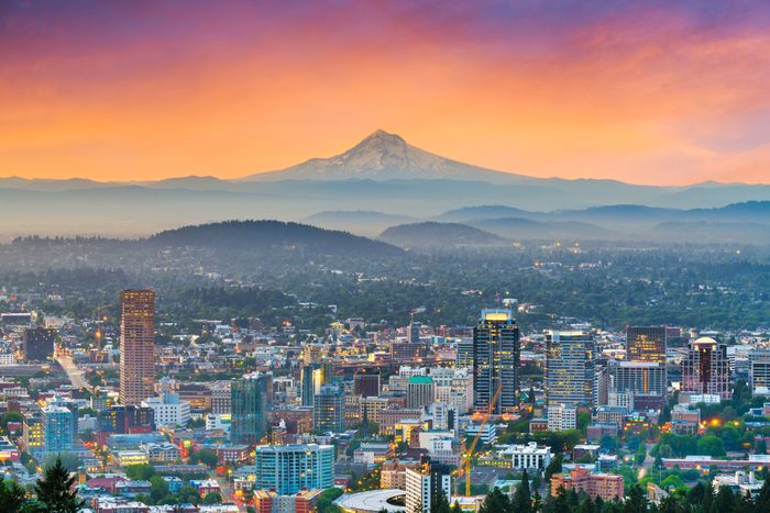 City buildings stand illuminated, overlooked by a distant snow-capped mountain, under a vibrant orange and purple sunset sky.