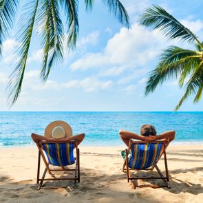 Two people recline in beach chairs, gazing at the sea under palm trees, with a clear blue sky above.
