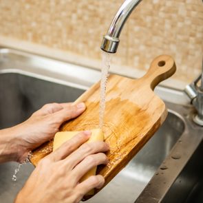 Hands scrub cutting board under a running faucet in a kitchen sink.