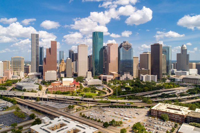 Skyscrapers stand tall under a bright blue sky with scattered clouds, surrounded by roads, greenery, and parking lots in an urban setting.