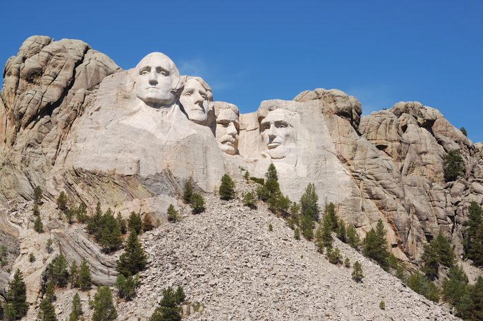 Giant stone faces carved on a mountain, surrounded by rocky terrain and scattered trees under a clear blue sky.