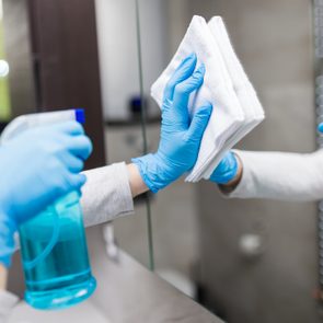 Gloved hands clean a mirror with a cloth and spray bottle in a bathroom setting.