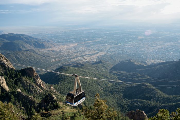 A cable car glides over a forested mountain range, with a vast cityscape and clear sky in the background.