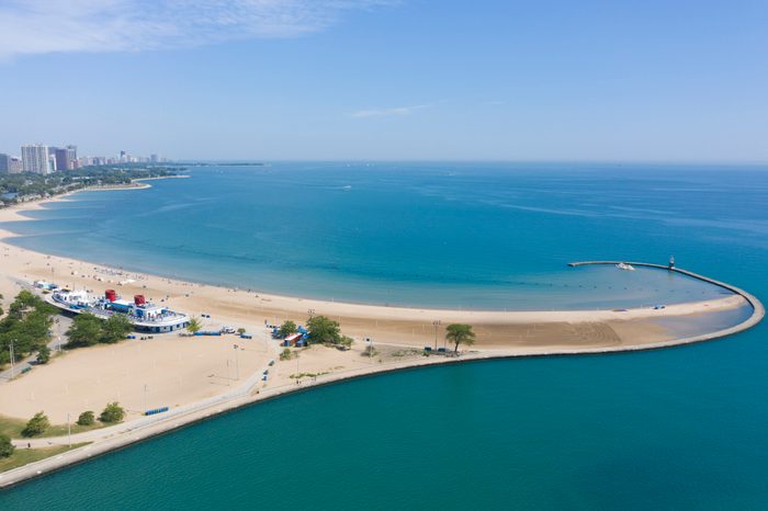 Beach curves along vibrant coastline, bordered by calm blue water, with distant city skyline under clear blue sky.