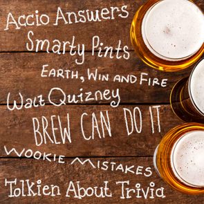 Beer glasses resting on a wooden table with playful quiz team names written in white chalk-like text, creating a playful trivia atmosphere.
