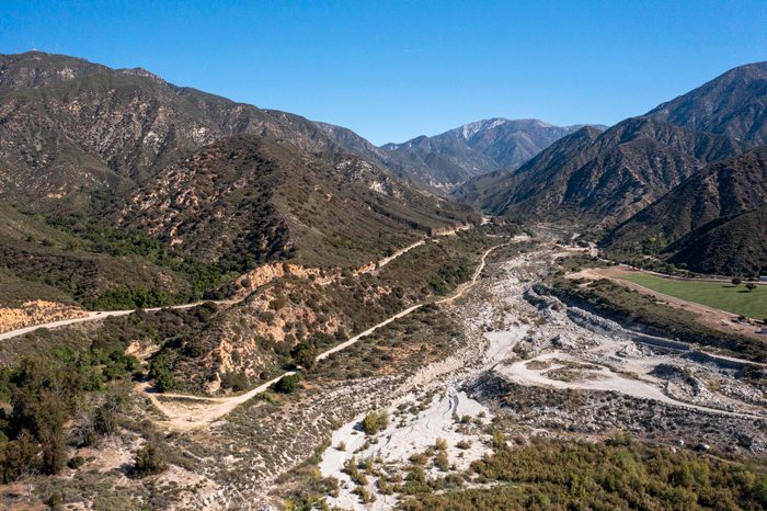 Mountains surround a winding road through a dry, rocky riverbed in a green valley under a clear blue sky.