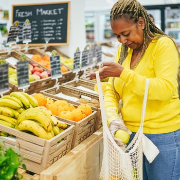 Woman selecting produce, placing fruits into a mesh bag, in a grocery store with various fruits displayed in wooden crates. A "Fresh Market" sign is present.