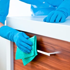 Gloved hand cleans a wooden cabinet handle with a green cloth in a bright, modern kitchen setting.