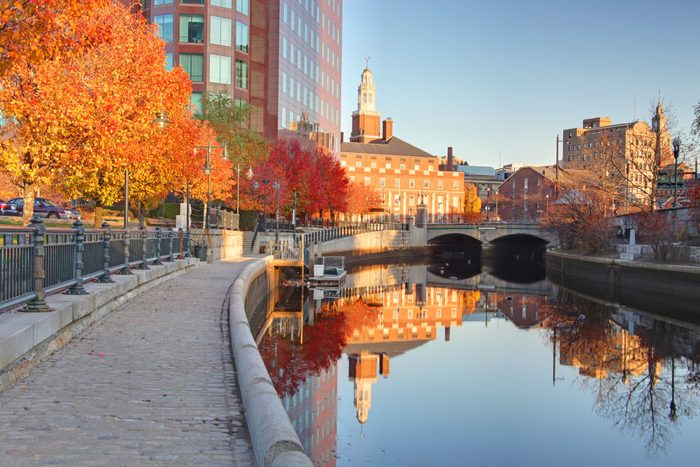 Tree-lined cobblestone path follows a calm river, reflecting colorful autumn foliage and buildings, with a bridge connecting a cityscape under a clear sky.
