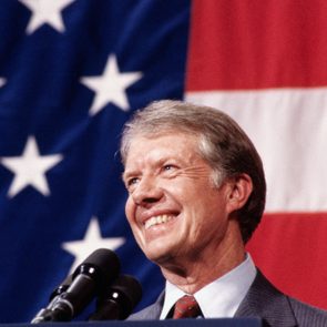 Smiling man stands behind microphones, against a backdrop of the American flag with stars and stripes.