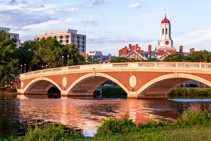 Arch bridge reflects on calm river; stately buildings with red roofs and spires in background; lush greenery surrounds. Text: 