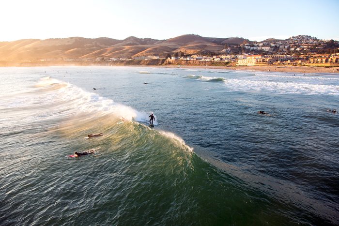 Surfer rides wave; others paddle nearby; town and hills overlook sunny beach.