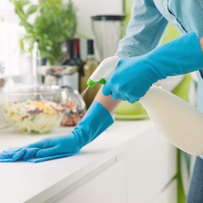 Gloved hands spray and wipe a countertop in a bright kitchen, with vegetables and jars in the background.