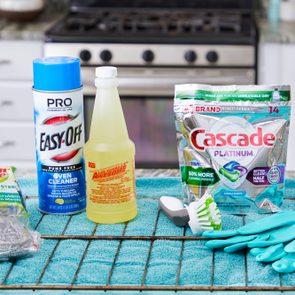 Cleaning products displayed on a kitchen counter include oven cleaner, detergent, scrubbing pads, gloves, and a brush, set against a stove backdrop.