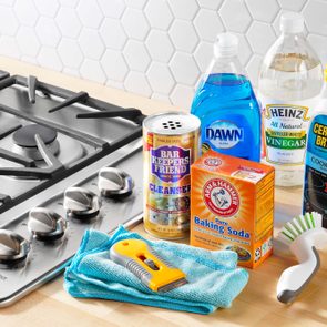 Cleaning supplies arranged. Products include Bar Keepers Friend, Dawn, vinegar, baking soda, Cerama Bryte, and a brush beside a gas stovetop on a kitchen counter.