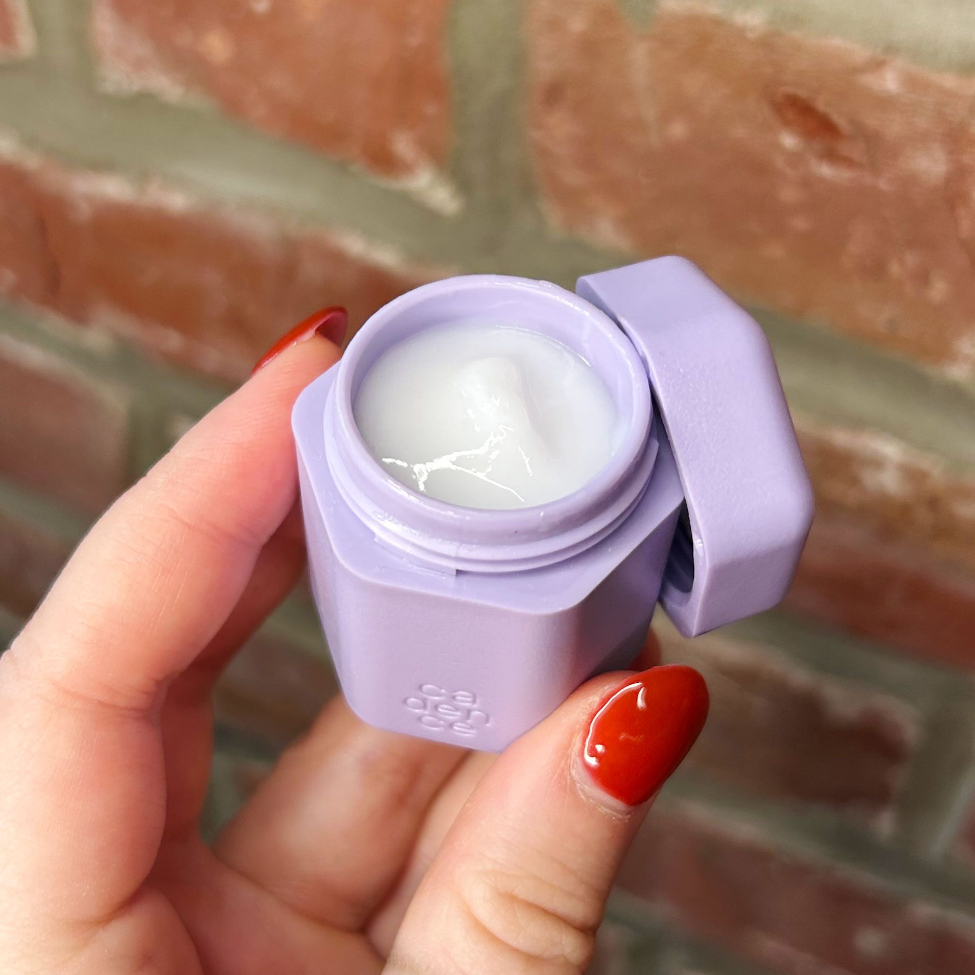 A purple container, held open, reveals white cream inside against a brick wall background.