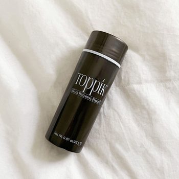 A black cylindrical bottle lies on a white fabric surface, labeled "Toppik Hair Building Fibers" and "Net Wt. 0.87 oz/25 g".