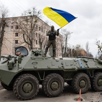 Armored vehicle, soldier waving Ukrainian flag; urban street with bare trees and building backdrop.