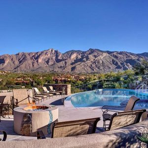 Pool reflecting sunlight, surrounded by lounge chairs. Mountains rise in the background under a clear blue sky, creating a serene outdoor setting.