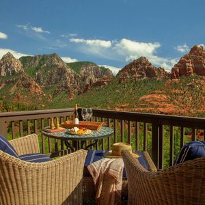 A table holds wine and snacks on a balcony, surrounded by wicker chairs, overlooking red rock formations under a clear blue sky.
