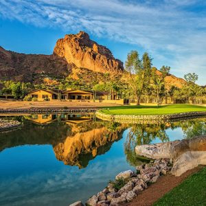 Brown mountain reflecting in the pond, surrounded by vibrant green grass and trees, with buildings nestled at its base under a clear blue sky.
